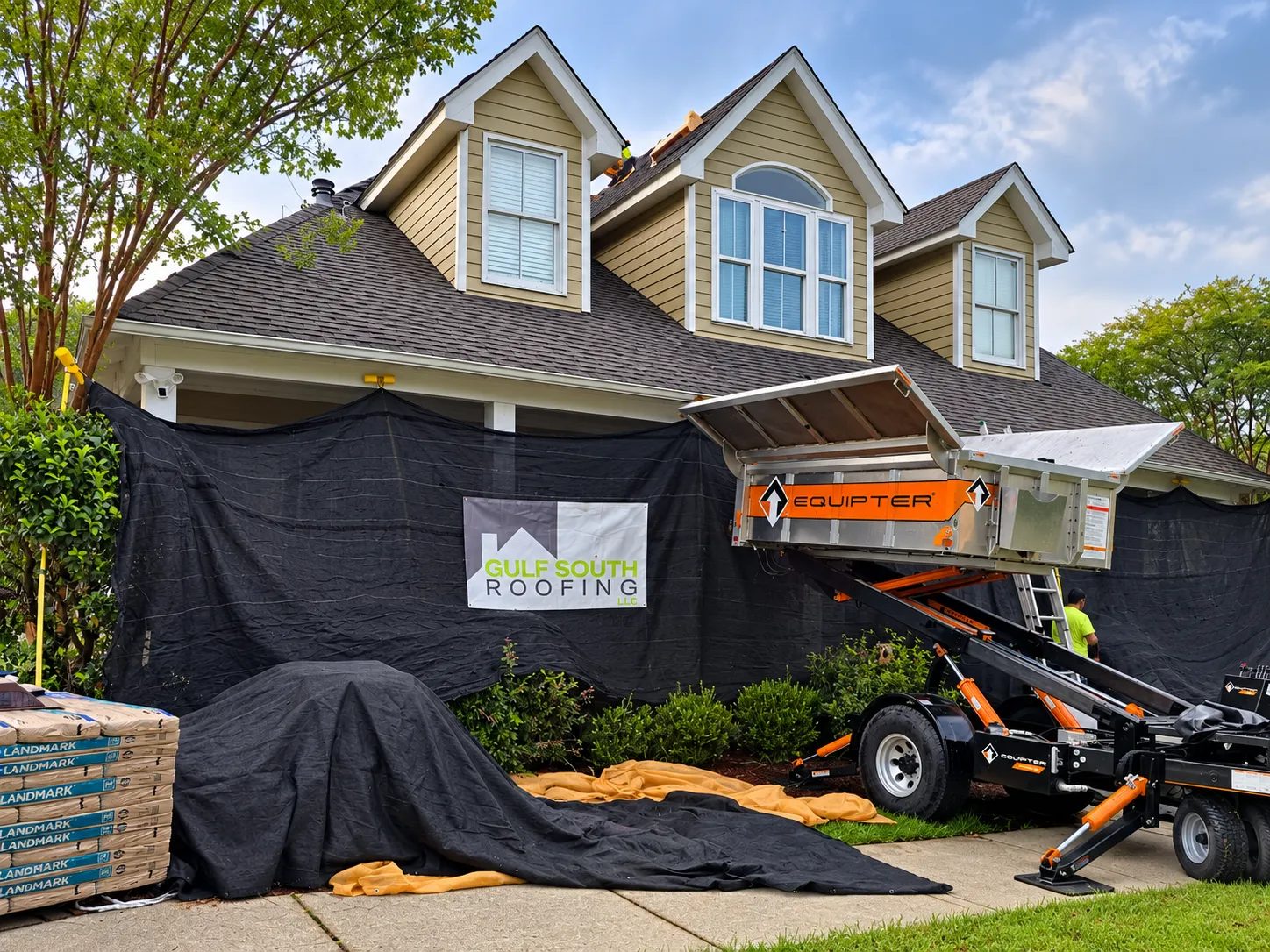 Gulf South Roofing crew tearing off an old shingle roof on a Gulf Coast home, the house wrapped in black debris netting, an Equipter lift catching the old shingles and new bundles stacked on the driveway