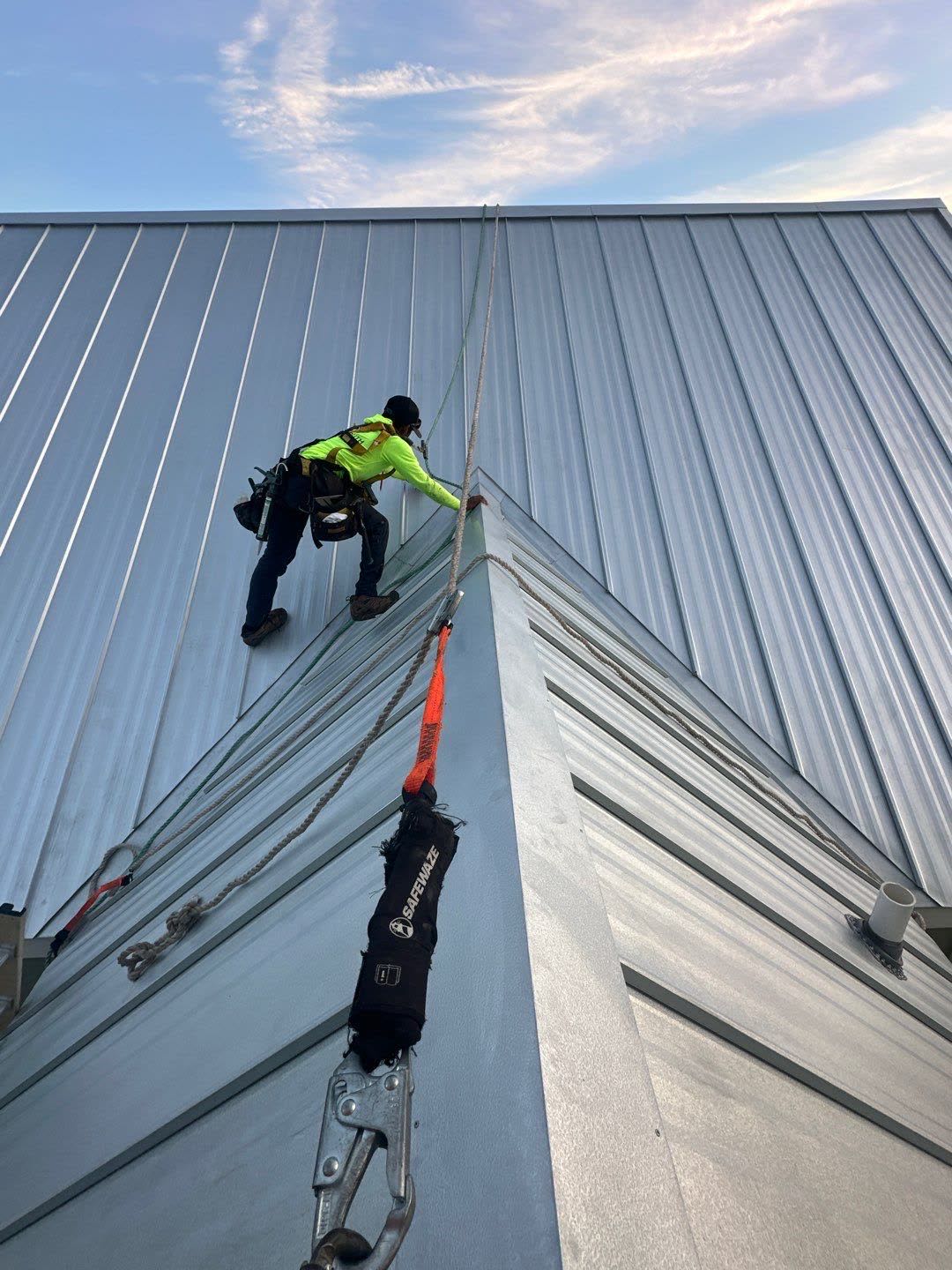 Gulf South Roofing crew member in high-visibility gear, safety-tethered while climbing a steep standing seam metal roof
