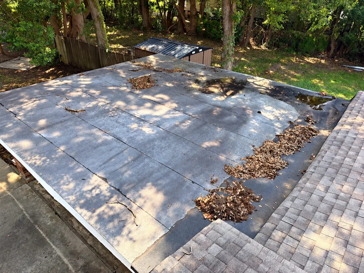 Leaves, debris and standing water on a worn low-slope roof section beside a shingle field on a Gulf Coast home: what bad drainage looks like