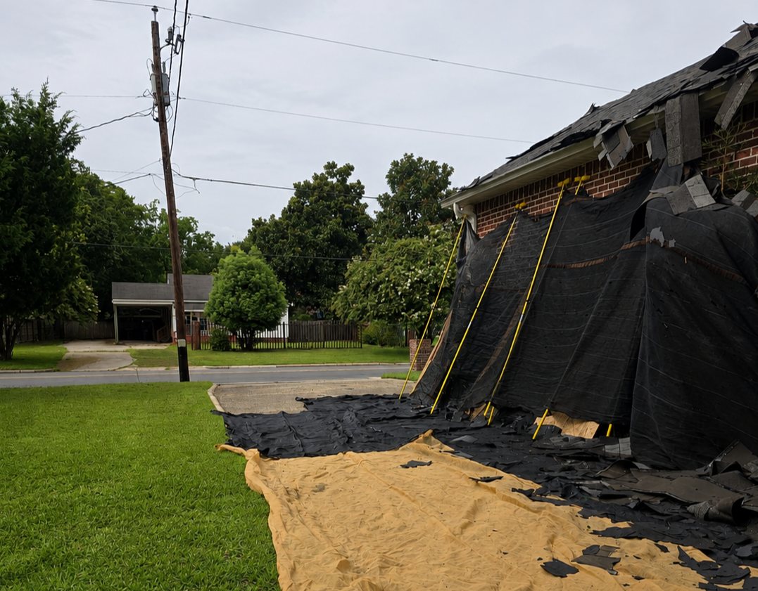 Debris netting hung from the eave and tarps spread on the lawn to catch tear-off debris during a shingle replacement